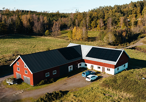 Röd lantgårdsbyggnad med solpaneler installerade av ZolarTech på taket, belägen i ett naturskönt område med öppna fält och skog i bakgrunden.